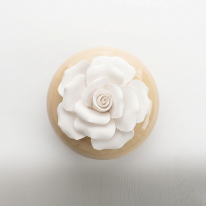 A round beige ceramic or stone vessel with a large sculpted white flower on top, photographed from above against a clean white background.