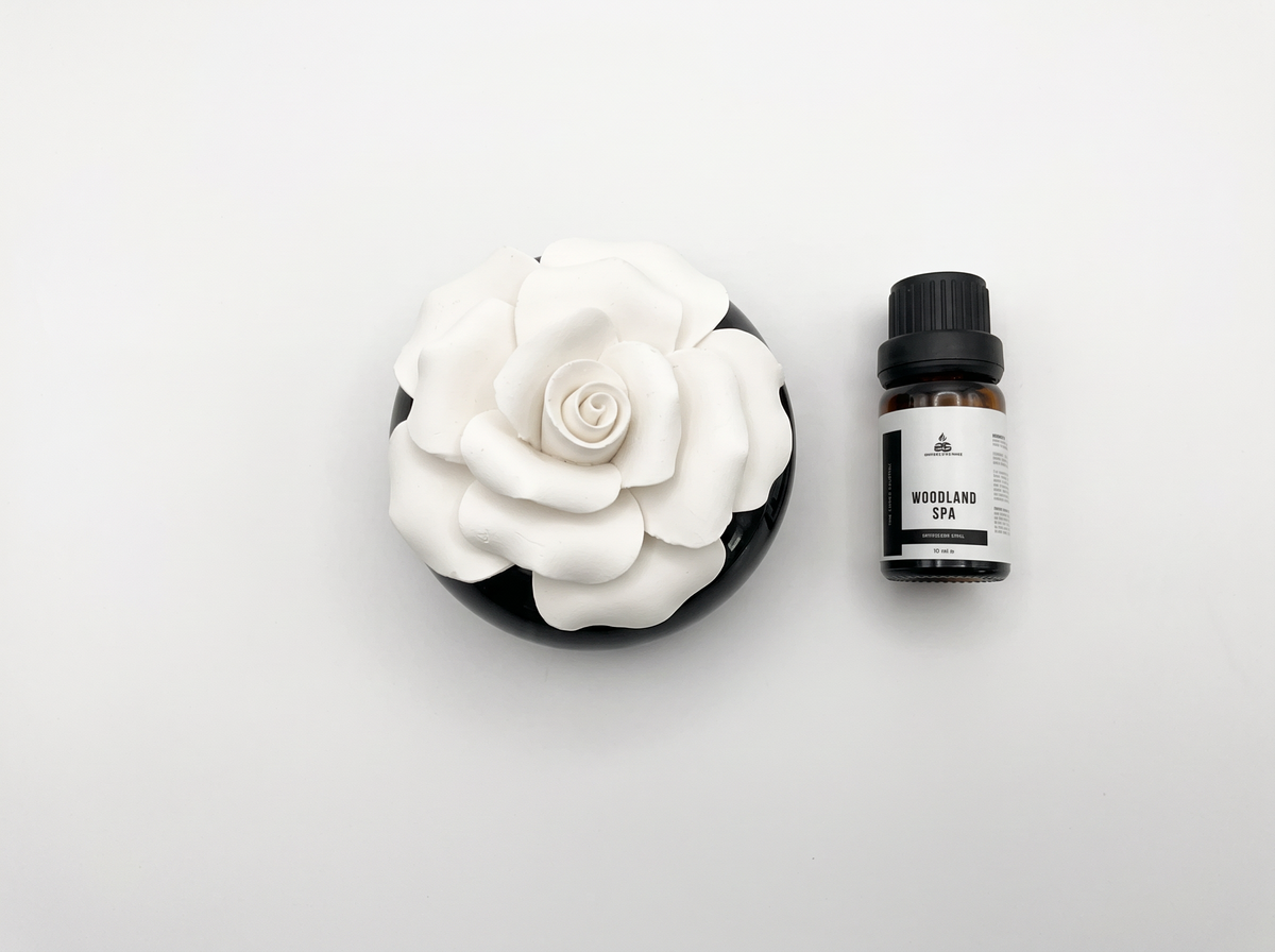 A white ceramic rose diffuser in a round black dish next to a small amber bottle labeled 'Woodland Spa' on a clean white background.