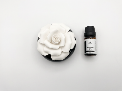 A white ceramic rose diffuser in a round black dish next to a small amber bottle labeled 'Woodland Spa' on a clean white background.