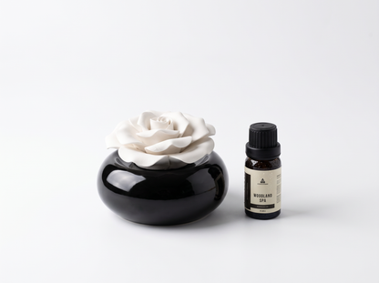 A black ceramic diffuser bowl with a white rose-shaped stone diffuser, shown next to a small dark amber bottle labeled 'Woodland Spa' on a clean white background.
