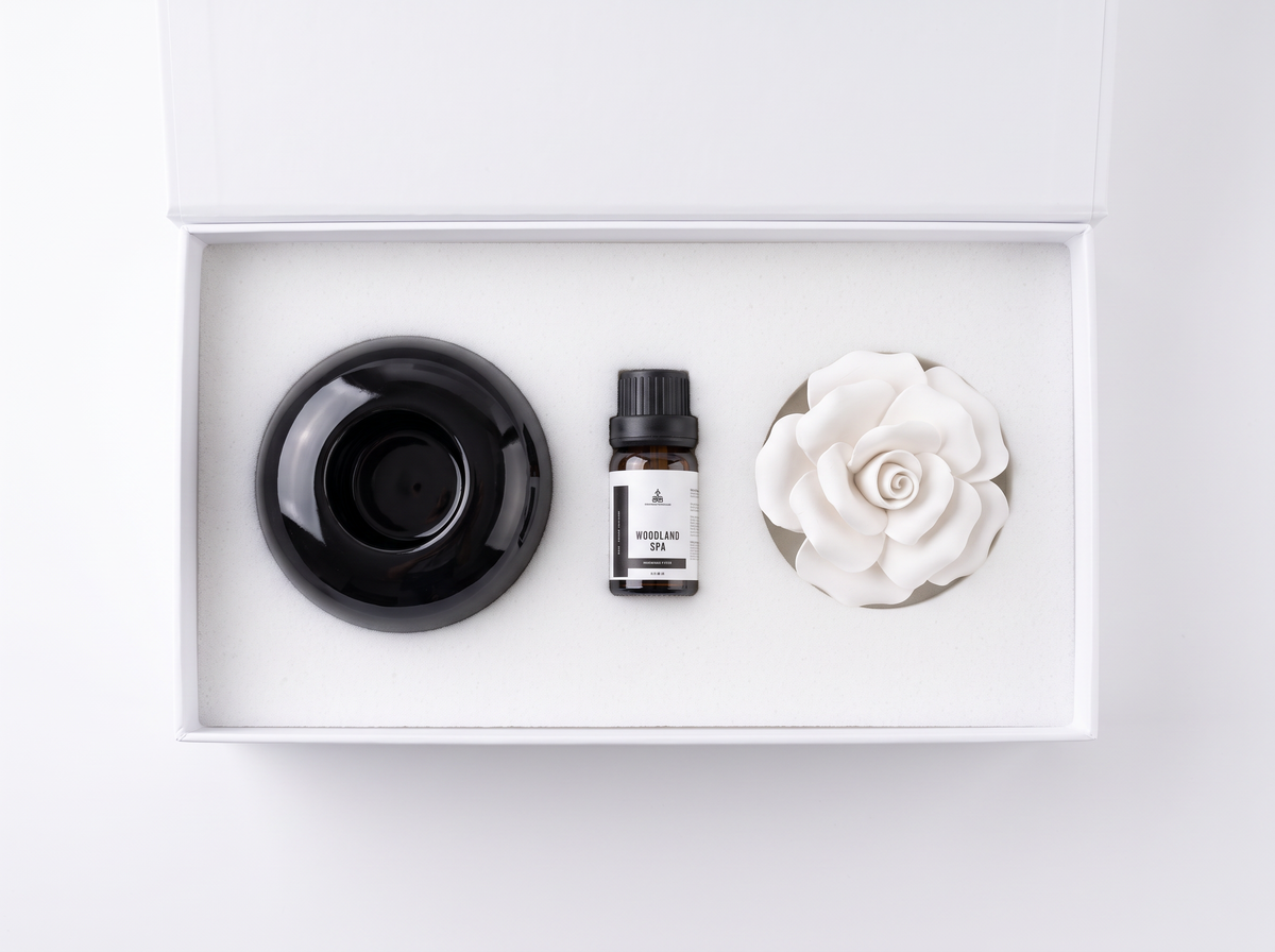 A diffuser gift set in a white box, featuring a black ceramic diffuser cup, a small bottle of fragrance oil, and a white ceramic rose-shaped diffuser.