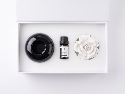 A diffuser gift set in a white box, featuring a black ceramic diffuser cup, a small bottle of fragrance oil, and a white ceramic rose-shaped diffuser.