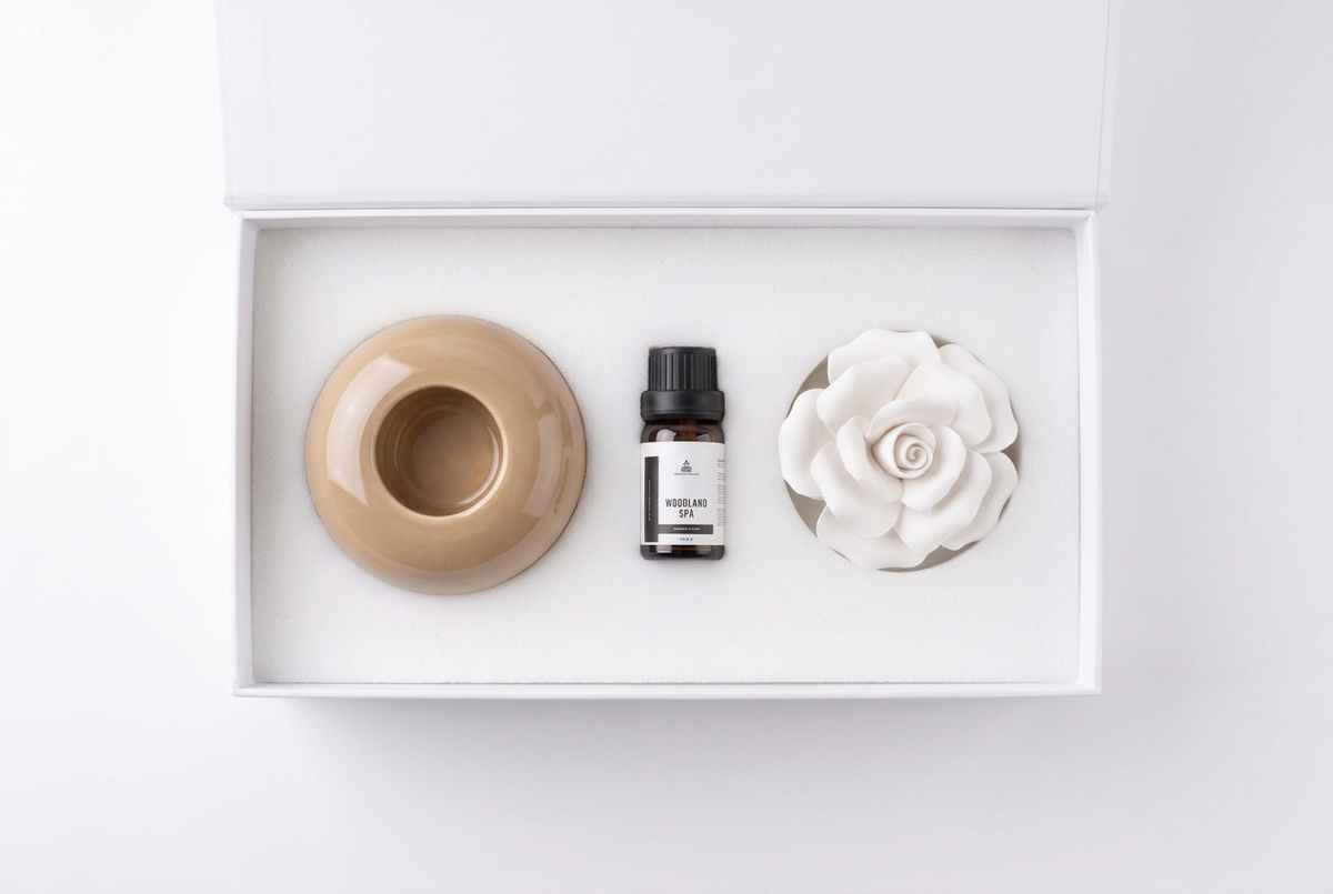 A minimalist gift set in a white box containing a beige ceramic bowl, a small amber bottle with a black cap and white label, and a white ceramic rose-shaped diffuser, photographed from above on a white background.