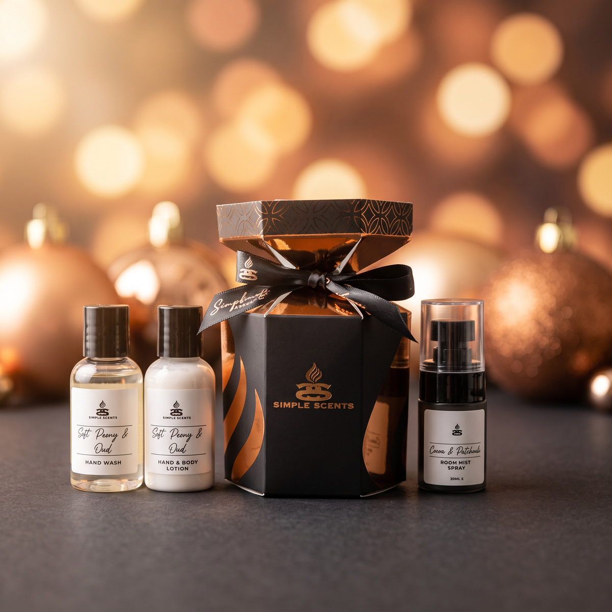 A gift set of Simple Scents home fragrance products arranged on a dark surface with a warm, blurred holiday bokeh background. The set includes a large black hexagonal box with a black ribbon, a small clear bottle of hand wash, a small white bottle of lotion, and a small black-and-white room spray bottle.
