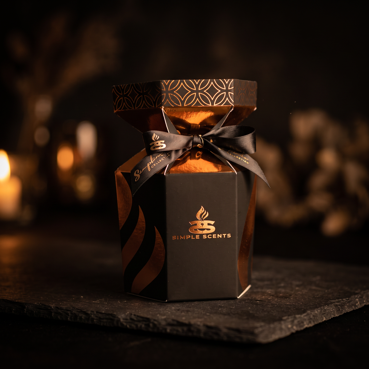 A hexagonal black gift box for Simple Scents, wrapped in a black ribbon and bow, set on a dark slate surface with a warm, blurred background.