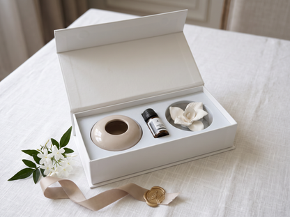 A white gift box opened on a white tablecloth, containing a beige ceramic diffuser, a small dark bottle, and a white flower-shaped diffuser insert. A beige ribbon and gold seal are placed beside the box, with soft neutral background blur behind it.