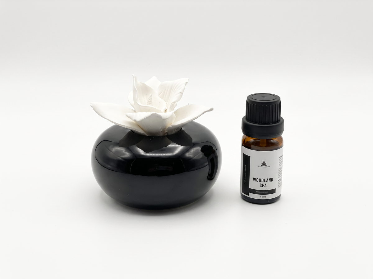 A small amber glass bottle with a black cap and white label next to a glossy black stone-like diffuser holder with a white flower-shaped top, on a light gray background.