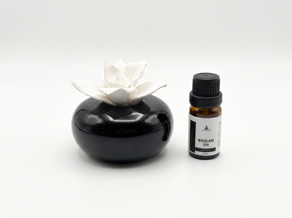 A small amber glass bottle with a black cap and white label next to a glossy black stone-like diffuser holder with a white flower-shaped top, on a light gray background.