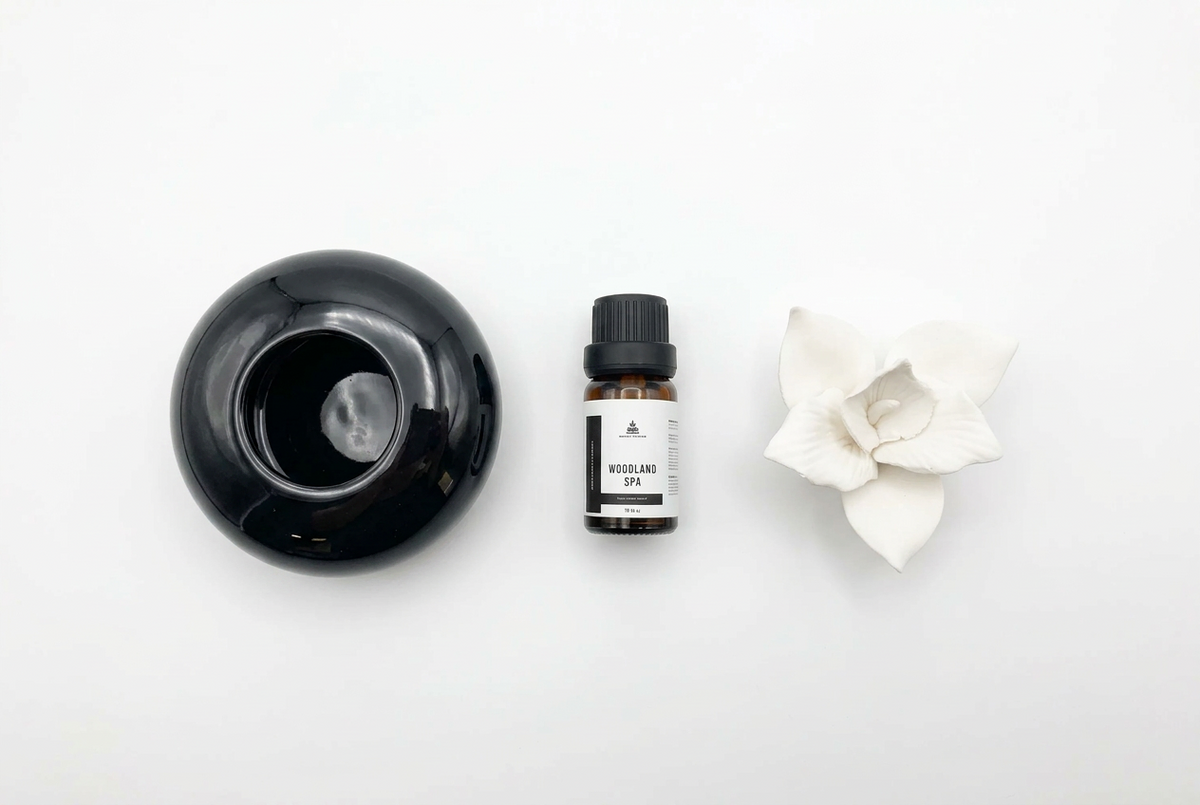 A small bottle of essential oil with a black cap and white label, placed between a glossy black diffuser bowl and a white flower on a clean white background.