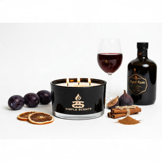 A black scented candle in a clear glass jar with three lit wicks and visible flames, branded Simple Scents. The candle is styled on a white background with dried orange slices, plums, a glass of red wine, a bottle of aged rum, cinnamon sticks, and figs, suggesting a warm spice or fruit fragrance.