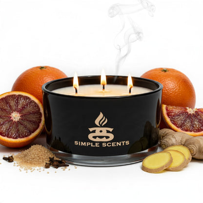 Lit Citrus Embers three-wick candle in a gloss-black jar, surrounded by blood oranges, brown sugar, cloves and sliced ginger.