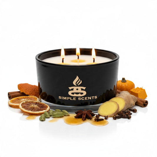 Lit Pumpkin Vale three-wick candle in a gloss-black jar, surrounded by pumpkin, dried orange, ginger, cardamom and warming spices