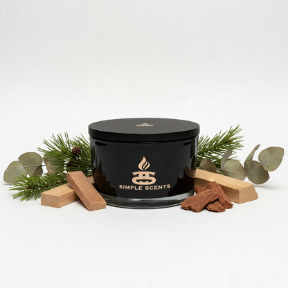 A black glass jar candle with a black lid and gold 'SIMPLE SCENTS' logo, styled with pine branches, eucalyptus leaves, small wooden blocks, and cinnamon sticks on a white background.