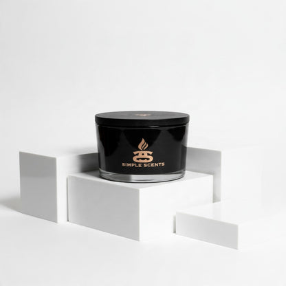 A black jar candle with a matching lid sits on a white geometric pedestal. The front of the jar is black with a gold logo and the words 'SIMPLE SCENTS'.