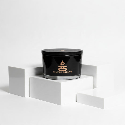 A black jar candle with a matching lid sits on a white geometric pedestal. The front of the jar is black with a gold logo and the words 'SIMPLE SCENTS'.