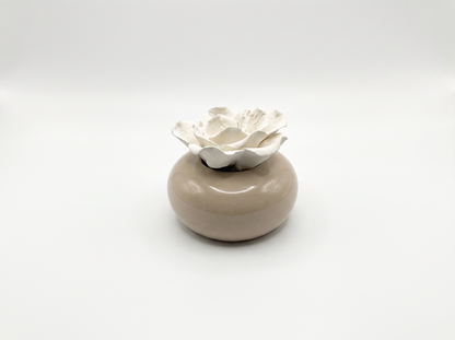 A small ceramic container with a smooth, rounded beige base and a white sculpted flower-shaped diffuser stone, set against a seamless light grey studio background.