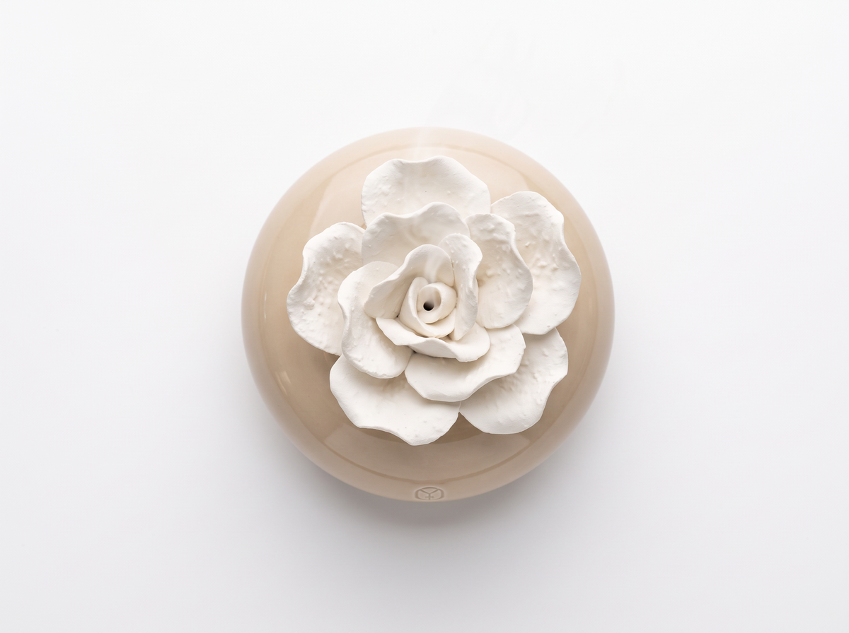 A round beige ceramic dish or planter with a large white sculpted flower on top, photographed from above on a clean white background.