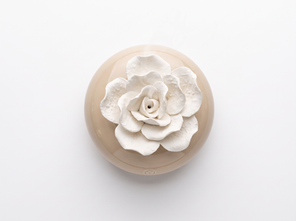 A round beige ceramic dish or planter with a large white sculpted flower on top, photographed from above on a clean white background.